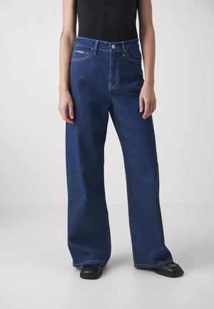 Personne portant un jean large en denim bleu foncé, un haut noir et des chaussures noires, debout devant un fond clair uni.
