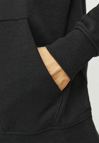 Crna dukserica s prednjim džepom, rebrastim manžetama i glatkom teksturom. Detalji uključuju šavove uz rubove i ležeran kroj.