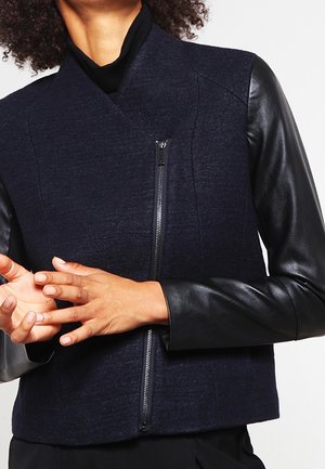 Femme portant une veste bleu marine avec des manches en cuir noir et une fermeture éclair asymétrique à l'avant, mains jointes au niveau de la taille.