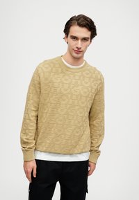 Μπεζ πουλόβερ με υφή λογότυπου, στρογγυλή λαιμόκοψη και ριγέ μανίκια. Φορεμένο πάνω από άσπρο πουκάμισο, συνδυασμένο με μαύρο παντελόνι.