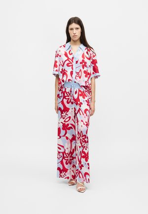 Woman wearing loose, short-sleeve button-up shirt and wide-leg pants with red and light blue floral patterns, standing against a white background.