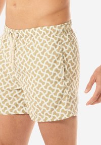 Costume da bagno beige con un motivo a onde bianche, vita elastica e lacci. Tasca laterale visibile, tessuto dalla superficie liscia.