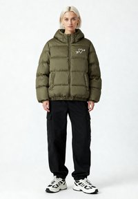 Doudoune vert olive avec capuche, ornée d'un motif brodé de cœur et flèche sur la poitrine. Portée avec un pantalon cargo noir et des baskets.