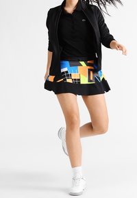 Černé polotriko s límečkem, doplněné barevnou plisovanou sukni s geometrickými tvary a černým pasem. Bílé tenisky.