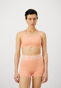 Lys ferskenfarget sportsbh med racerback-design og hvit Nike-logo, kombinert med matchende høyt midjete shorts med "NIKE PRO"-livbånd.