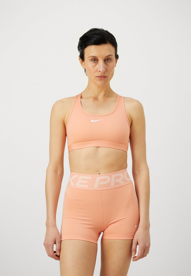 Lys ferskenfarget sportsbh med racerback-design og hvit Nike-logo, kombinert med matchende høyt midjete shorts med "NIKE PRO"-livbånd.
