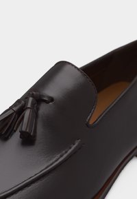 Mocassin en cuir brun foncé avec des pampilles décoratives et des coutures visibles sur un fond blanc épuré.