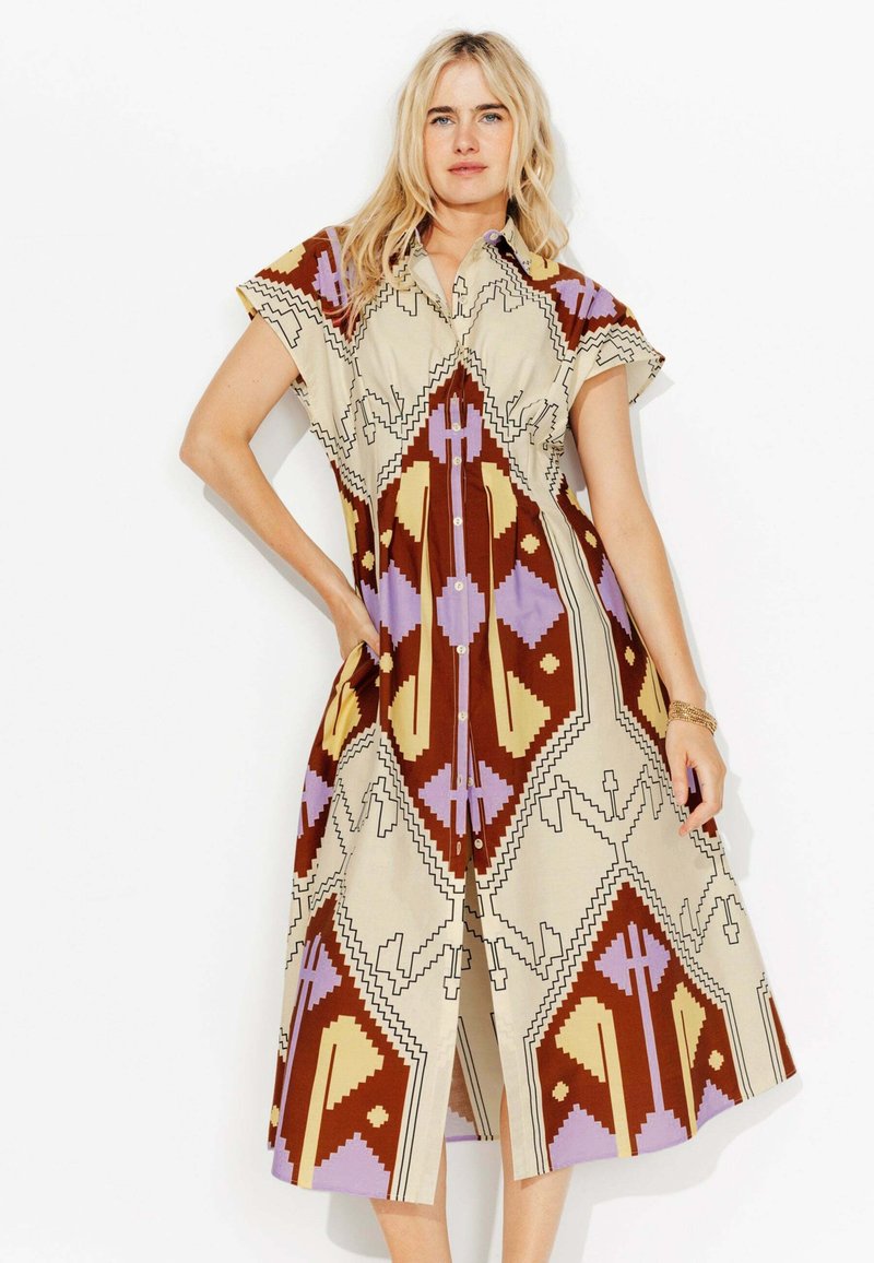 Robe en button-up de couleur taupe avec de grands motifs géométriques en brun, lavande et jaune. Manches courtes et poches latérales sont très présentes.