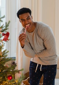 Grijs shirt met lange mouwen, marineblauwe pyjamabroek met zuurstokpatroon, houdt een roze gestreepte zuurstok vast, staand bij een kerstboom.