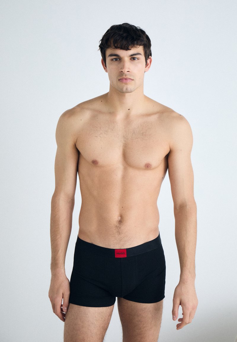 Schwarze Boxershorts mit weitem Bund und rotem Logo. Glatte Textur, taillierte Passform und kurze Beinlänge. Modell von vorne gezeigt.
