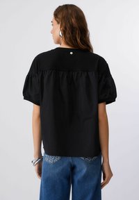 Črn top z nabranimi ovratniki in nabranim hrbtom. Ima okrogel izrez in majhen srebrn gumb na zgornjem delu hrbta.