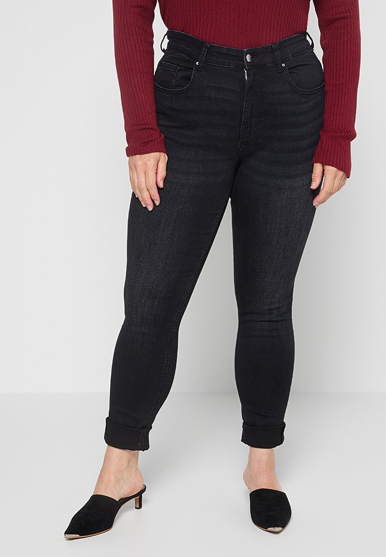 Schwarze, enge Jeans mit hoher Taille, Fronttaschen und einer leicht abgenutzten Textur. Kombiniert mit einem kastanienbraunen, gerippten Oberteil und schwarzen Pantoletten.