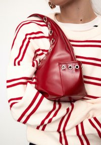 Borsa rossa in pelle con forma strutturata, caratterizzata da dettagli con occhielli argentati lungo la tracolla e una tasca laterale. Indossata sopra un maglione a righe.