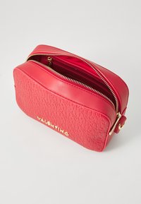 Trousse de maquillage en cuir rouge avec une surface texturée présentant des motifs de logo en relief. Accents en fermeture éclair dorée et ouverture zippée sur le dessus.