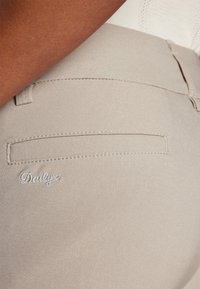 Pantalons beiges avec une texture lisse, dotés d'une poche arrière et d'un logo brodé "Daily" en blanc. Design minimaliste avec des lignes épurées.