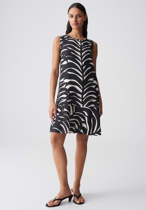 Donna con vestito senza maniche nero e bianco con stampa a zebra lungo fino al ginocchio con orlo arricciato e sandali neri con tacco su sfondo neutro.