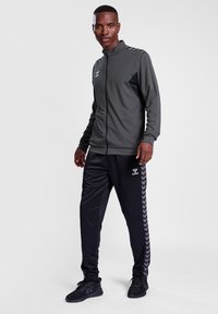 Sportief trainingspak met een grijze zip-jack met zwarte accenten en zwarte broek met een visgraatpatroon. Het materiaal lijkt glad en lichtgewicht.