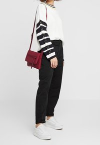 Bílý svetr s námořnicky modrými pruhovanými rukávy, černé kalhoty a bílé tenisky. Červená kabelka přes rameno se zipem. Ležérní outfit.