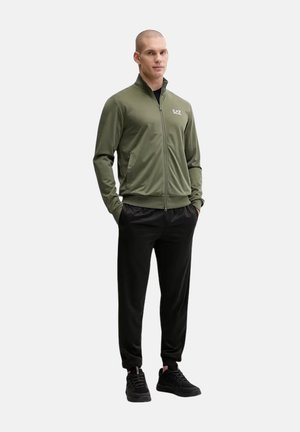 Uomo che indossa una giacca verde oliva con zip e pantaloni neri con sneakers nere, in piedi con le mani nelle tasche su sfondo semplice.