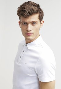 Weißes Polo-Shirt mit Kragen, Drei-Knopf-Leiste und dezentem Logo auf der Brust. Glatte Stoffstruktur, lockere Passform.