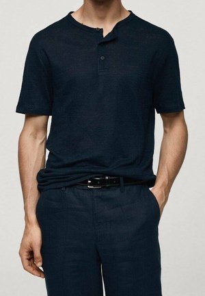 Mand iført en mørkeblå kortærmet henley-skjorte, stoppet ned i matchende marineblå bukser med sort bælte, hænder afslappet langs siderne.