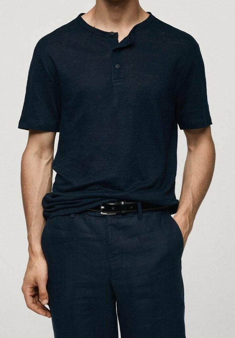 Homme portant un polo henley à manches courtes bleu marine foncé, rentré dans un pantalon bleu marine assorti avec une ceinture noire, les mains détendues le long du corps.