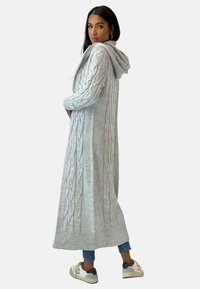 Lange grijze gebreide cardigan met een kabelpatroon en een capuchon, voorzien van ribgebreide manchetten en zoom. Gecombineerd met jeans en sneakers.