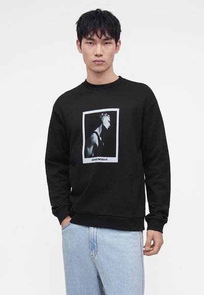 Jeune homme portant un sweat-shirt noir avec un motif de portrait monochrome et un jean bleu clair, debout avec une main dans la poche.