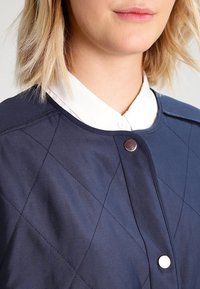 Detailní záběr na osobu v námořnické prošívané bundě se stříbrnými druky přes bílou košilovou košili.