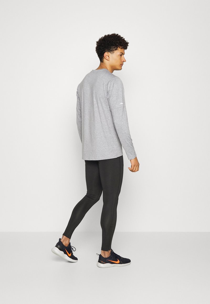 Joven caminando, vistiendo una camiseta de manga larga gris claro, leggings negros y zapatillas negras con el logotipo de Nike en naranja.