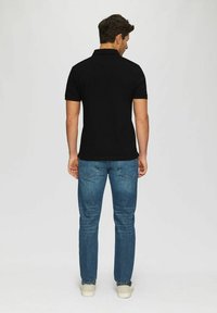 Schwarzes Polo-Shirt mit kurzen Ärmeln, kombiniert mit blauen Denim-Jeans. Der Stoff wirkt glatt und die Passform ist lässig. Legere Schuhe runden das Outfit ab.