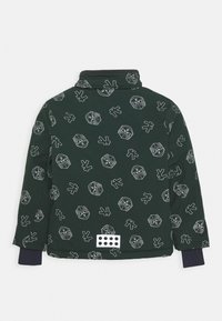 Zelený pulover s vysokým límcem, se vzory bílých abstraktních obličejů. Obsahuje obdélníkový bílý detail s černými tečkami na lemu.