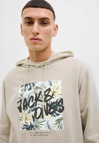 Beige hoodie med et grafisk print af tropiske planter og teksten "JACK & JONES." Lavet af blødt materiale med en kengurulomme og bindebånd i hætten.