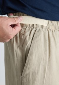 Pantaloni bej cu talie elastică, dintr-un material texturat, având buzunare laterale și o croială lejeră. O mână prinde talia pentru ajustare.