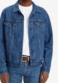 Veste en denim bleu avec deux poches poitrine, fermetures à bouton et un design texturé. Portée sur un t-shirt blanc et associée à un jean en denim.