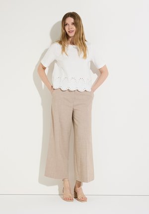 Frau mit langen Haaren trägt ein weißes Top mit kurzen Ärmeln und gewelltem Rand, beige weite Hosen und beige Sandalen mit Blockabsatz und Riemchen.