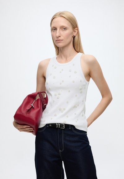 Femme aux cheveux blonds raides portant un haut blanc sans manches avec de petits détails floraux, un jean foncé, tenant un sac à main en cuir rouge sous le bras.