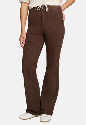 Vrouw draagt donkerbruine rechte broek met hoge taille en knoop- en ritssluiting, gecombineerd met witte schoenen en een crèmekleurige top.