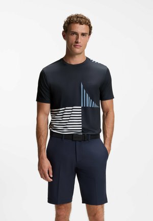 Mann trägt ein schwarzes T-Shirt mit blauen und weißen Streifen und marineblauen Shorts, steht mit einer Hand in der Tasche vor weißem Hintergrund.