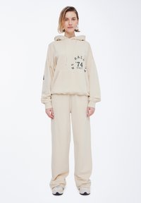 Sudadera beige y pantalones de chándal a juego, confeccionados en una tela suave, con una gran impresión de logo en el frontal y un bolsillo delantero. Diseño de corte holgado.