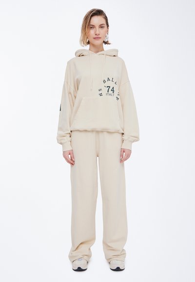 Beige hettegenser og matchende joggebukser laget av mykt stoff, med et stort logotrykk på fronten og en lomme foran. Løs passform.