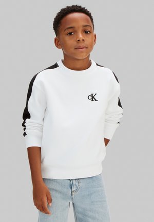 Junge trägt weißen Calvin Klein Pullover mit schwarzen Streifen an den Ärmeln und hellblaue Jeans, steht vor grauem Hintergrund.