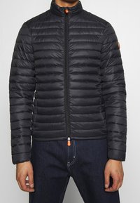 Giubbotto puffer nero con colletto alto, texture trapuntata, zip completa con tirante arancione e design aderente. Indossato sopra un paio di pantaloni di jeans.