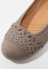 Ballerine en daim taupe avec une bande décorative perforée et cloutée sur l'avant, dotée d'une semelle intérieure rembourrée avec des trous de ventilation.