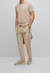 Polo beige avec un col noir et blanc, associé à un pantalon beige assorti. Des baskets aux teintes terre complètent l'ensemble.