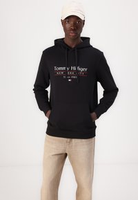 Schwarzer Kapuzenpullover mit Fronttasche; verfügt über weißen und roten "Tommy Hilfiger" Logo-Text. Kombiniert mit hellbeigen Jeans.