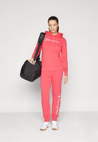 Felpa rossa in cotone con tasca frontale e logo bianco, abbinata a pantaloni jogger rossi con dettagli del logo. Borsa a tracolla nera, sneakers bianche.