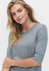 Blond kvinna med blå ögon som bär en grå kortärmad tröja, leende milt med armarna i kors mot en enfärgad vit bakgrund.