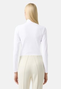 Weißes Langarmoberteil mit Stehkragen, ausgestattet mit Raglanärmeln und schmaler Passform. Kombiniert mit hochgeschnittenen cremefarbenen Hosen.