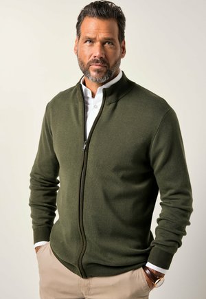 Man with beard wearing green zip-up sweater, white shirt, beige pants, and wristwatch, posing with hands in pockets against neutral background.
